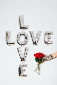 "Letter of Love: Redefining Relationships in Modern Times" 2 Silver balloons forming 'Love' with a hand holding red roses on a white background.