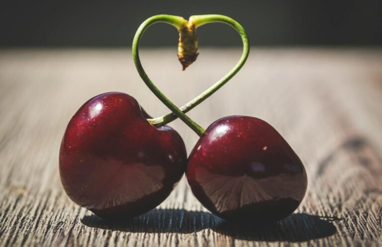 The 10 Amazing Benefits of Love for Health and Happiness (2025)happiness,mental health,benefit of love,Close-up of two ripe cherries with heart-shaped stems on a wooden table, symbolizing love.