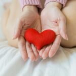 2025 Guide: Love – The Ultimate Secret Weapon for Your Wellness and Health.(Unrecognizable female sitting with bare legs on white sheet with small red heart in hands in light room in daytime)