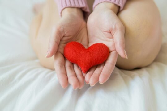 2025 Guide: Love – The Ultimate Secret Weapon for Your Wellness and Health.(Unrecognizable female sitting with bare legs on white sheet with small red heart in hands in light room in daytime)